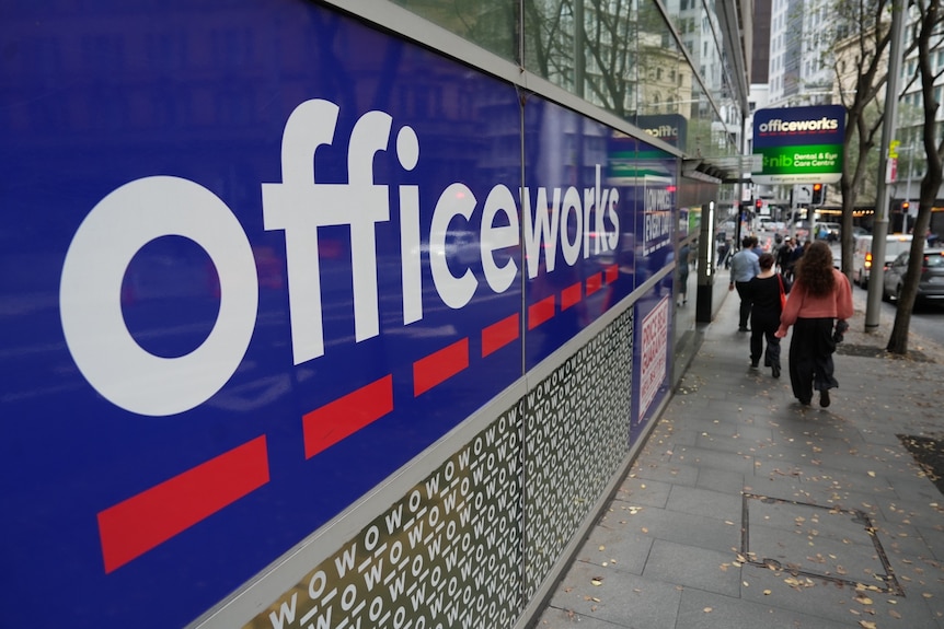 People walking outside on a street path past large Officeworks signage on windows of a building.