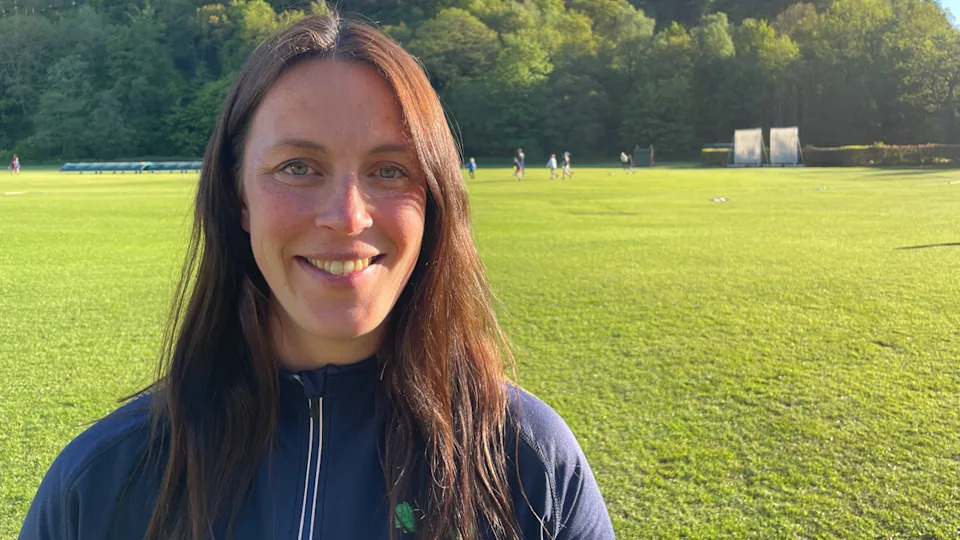 A woman wearing a navy top smiles at the camera. She is stood on a grass lawn, with a cricket pitch behind her.