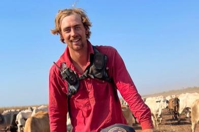 Jake, smiling, red hair, beard rides a bike with cattle behind him, wears red shirt, harness over it, blue sky.