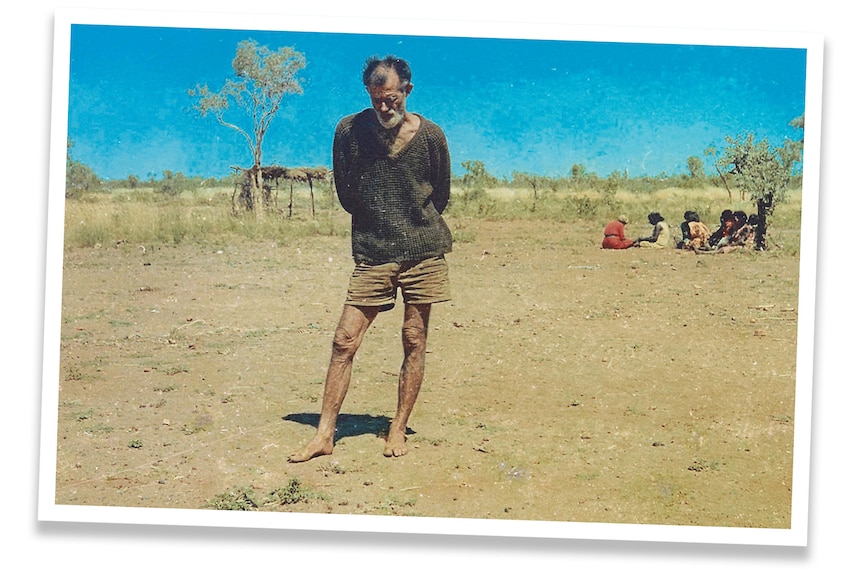 A man in a v-neck wool sweater and shorts stands barefoot, looking at the sand beneath his feet.