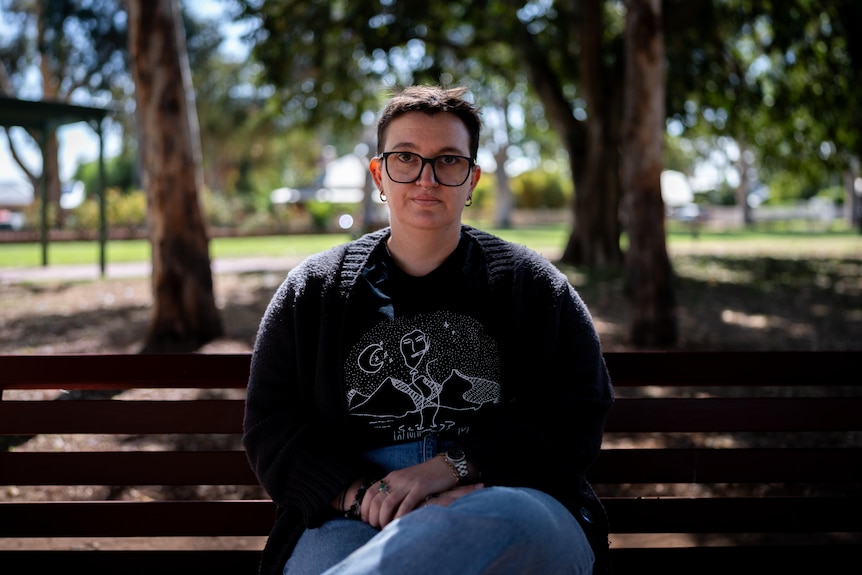 Le with glasses sits outdoors in a park. Is serious, short hair, dark top and jeans, on a bench.