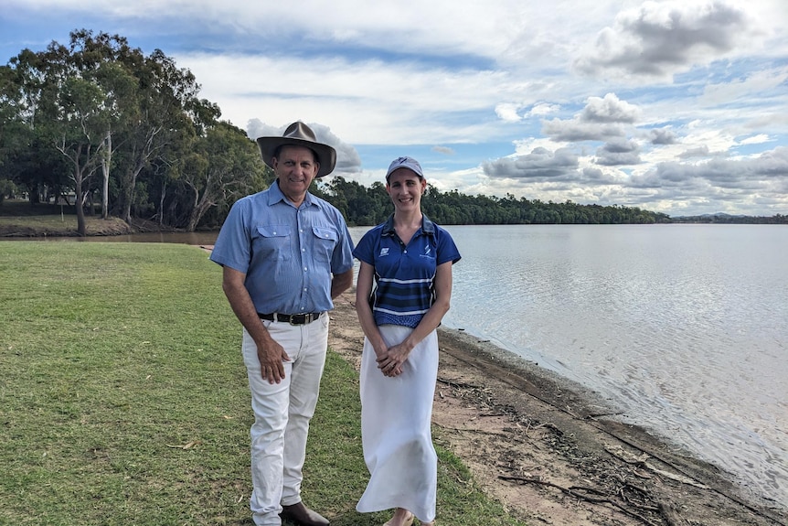 A man and woman smile as they stand on grass at the egde of a wide river lined by trees.