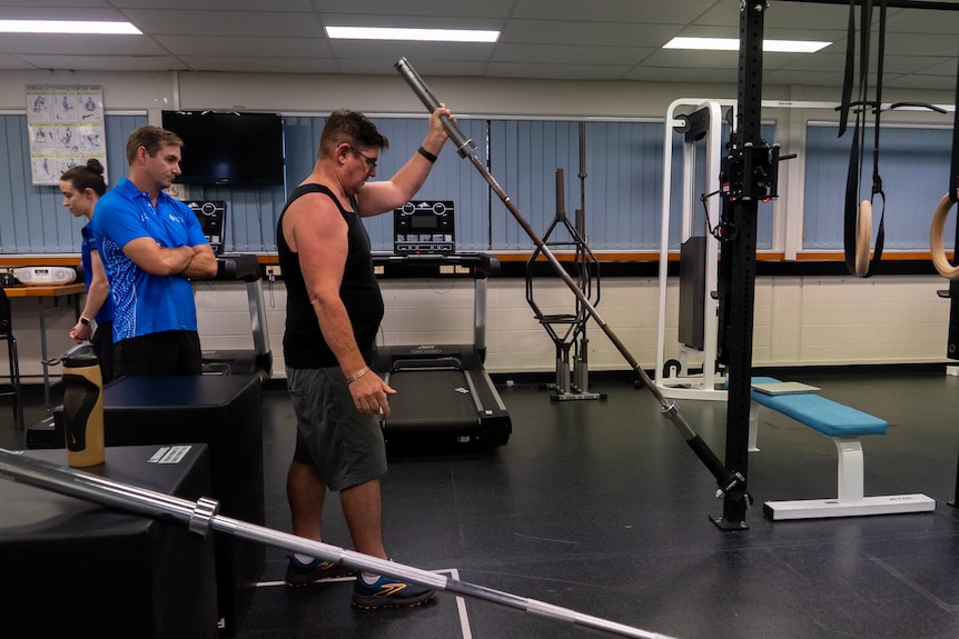 A man in a blue shirt watches a man in a black singlet pushing a weight bar.