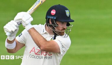 Ben Raine wearing a helmet with a Durham badge on it swooshes his bat high above his shoulder
