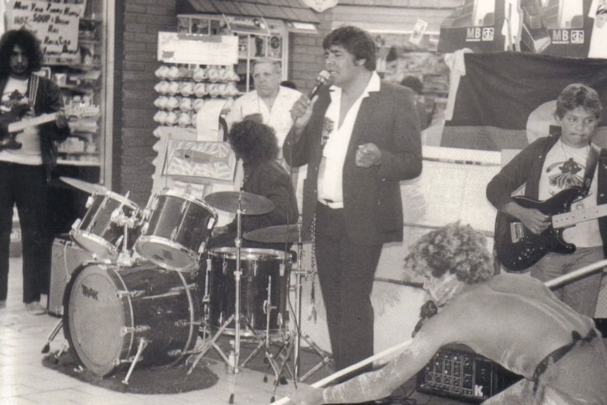 Roger Knox singing into a microphone next to a band.
