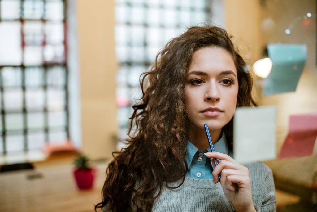 A focused woman holding a pen to her chin while looking at sticky notes on a glass board.