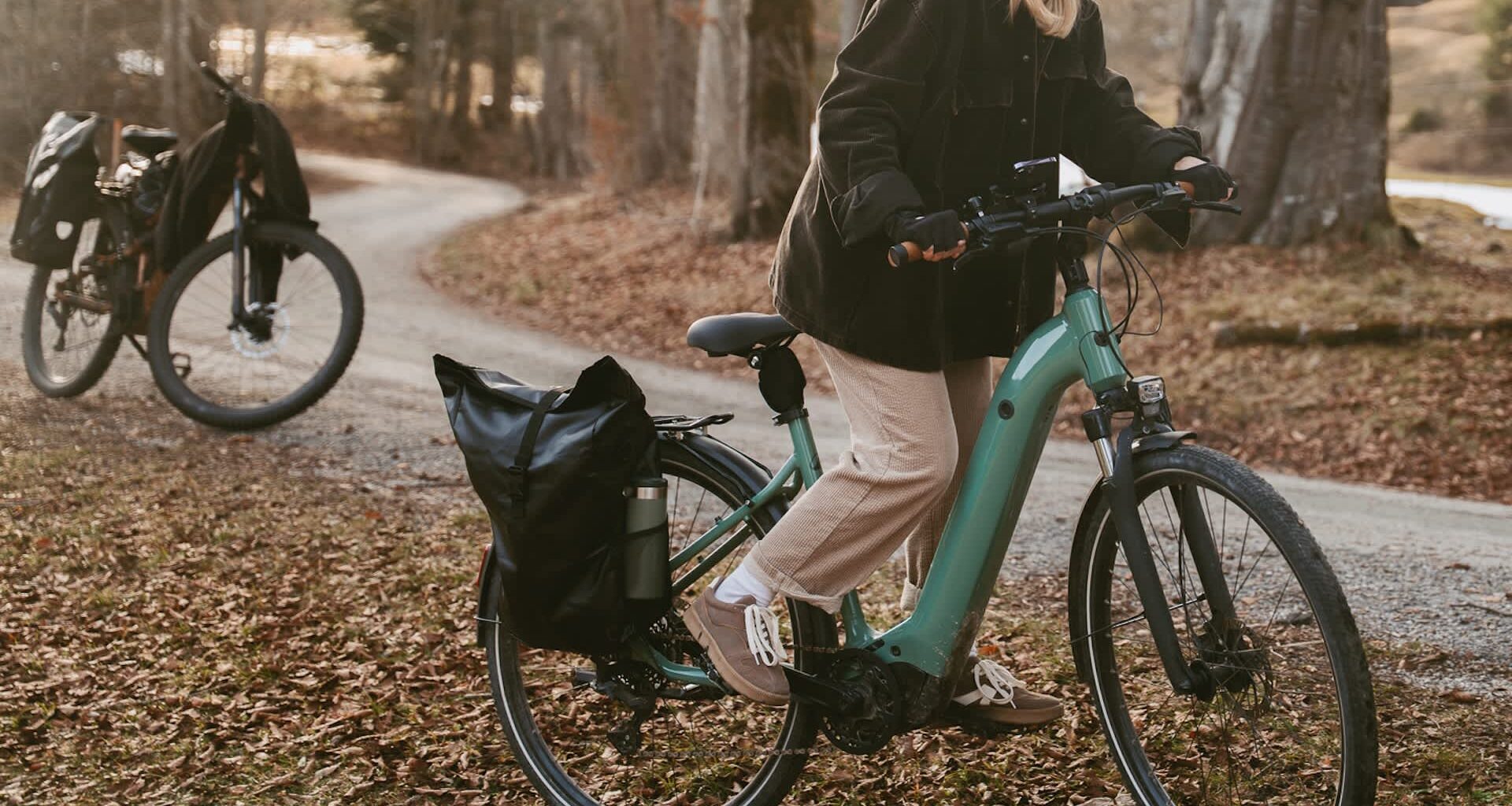 A woman in a black jacket rides a teal electric bike along a leaf-covered path with another bike in the background.
