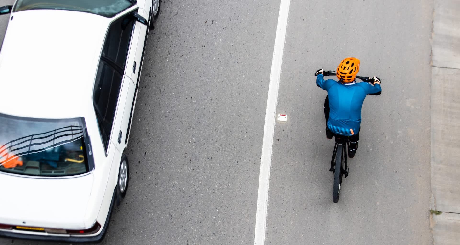 A cyclist in a blue shirt and orange helmet rides alongside a white car on a gray road.