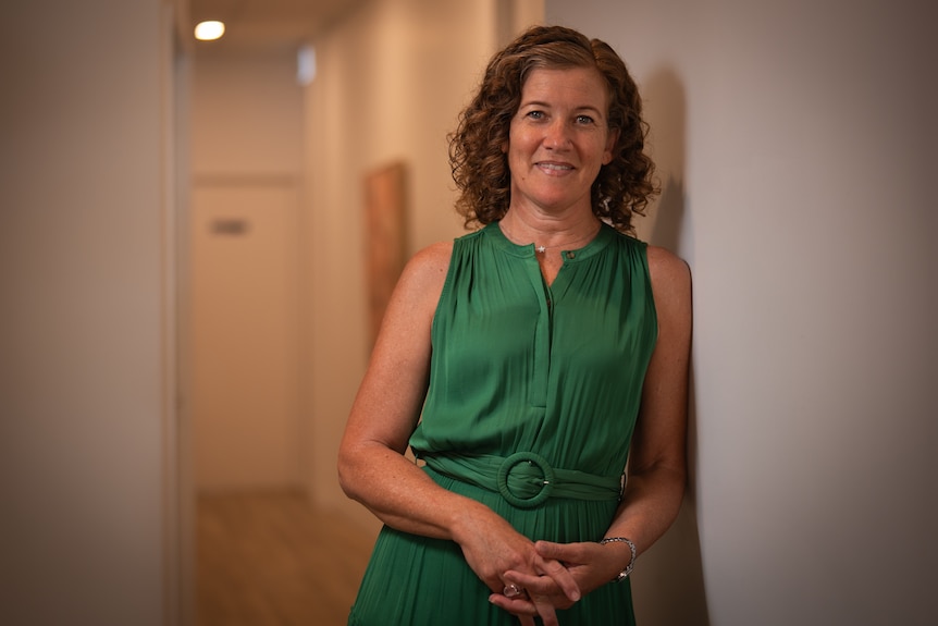 Woman in green dress smiles at camera while leaning on wall in clinic.