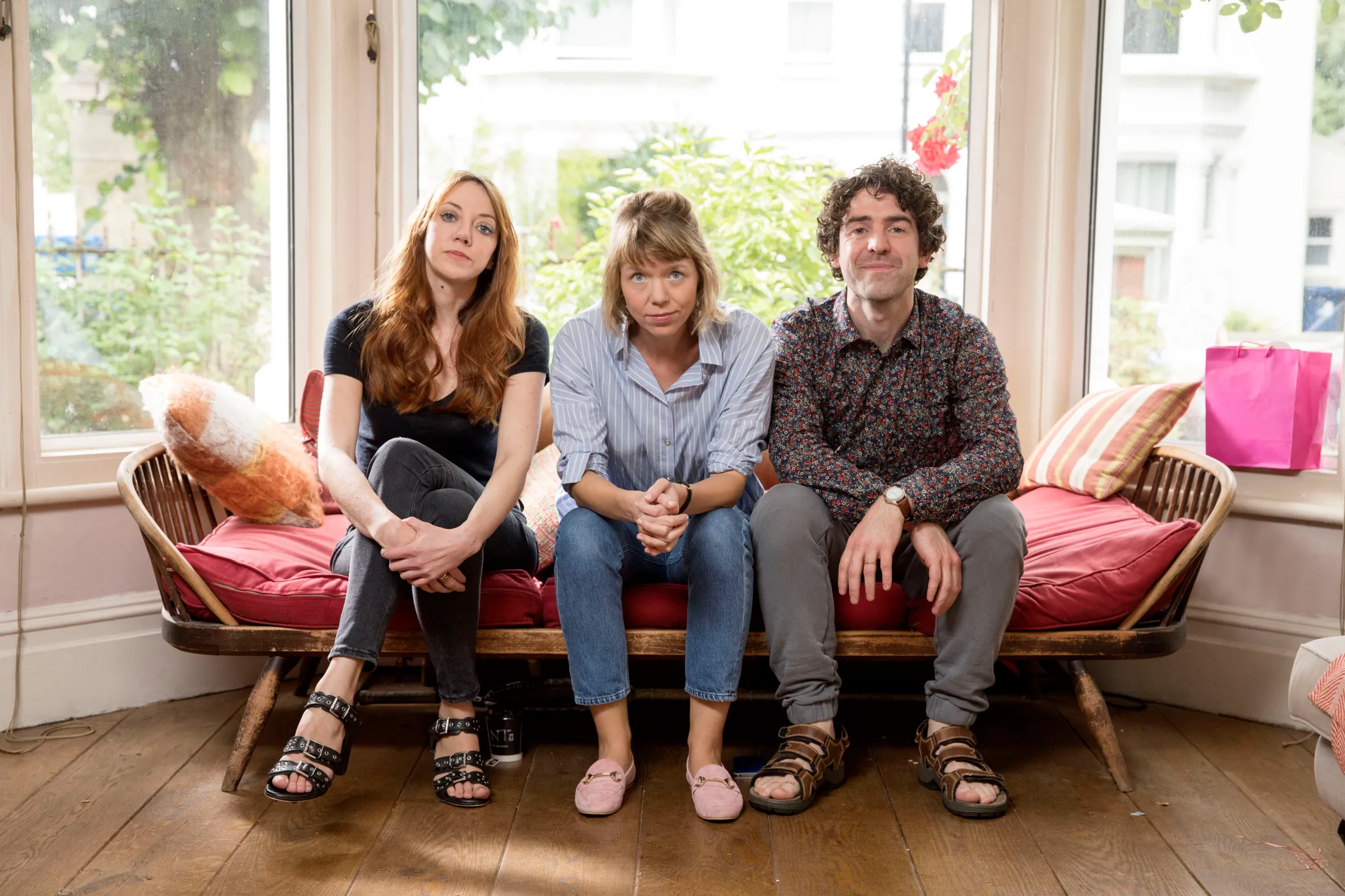 Diane Morgan, Anna Maxwell-Martin, and Paul Ready sitting on a wooden bench with red cushions.