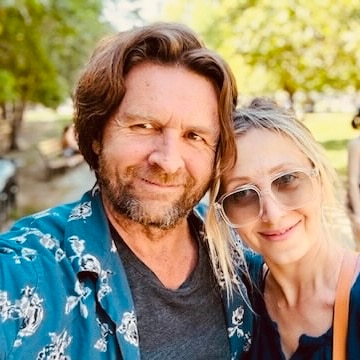 Man wearing a blue patterned shirt over a grey T-shirt, his arm around his wife who wears sunglasses and green top