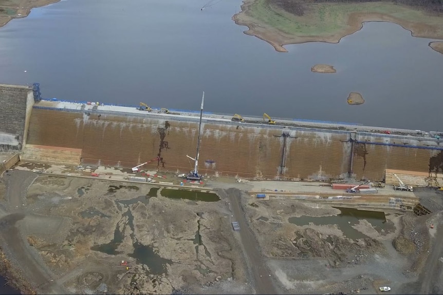 Aerial shot of construction work on a dam wall, looking across the spillway and upstream.