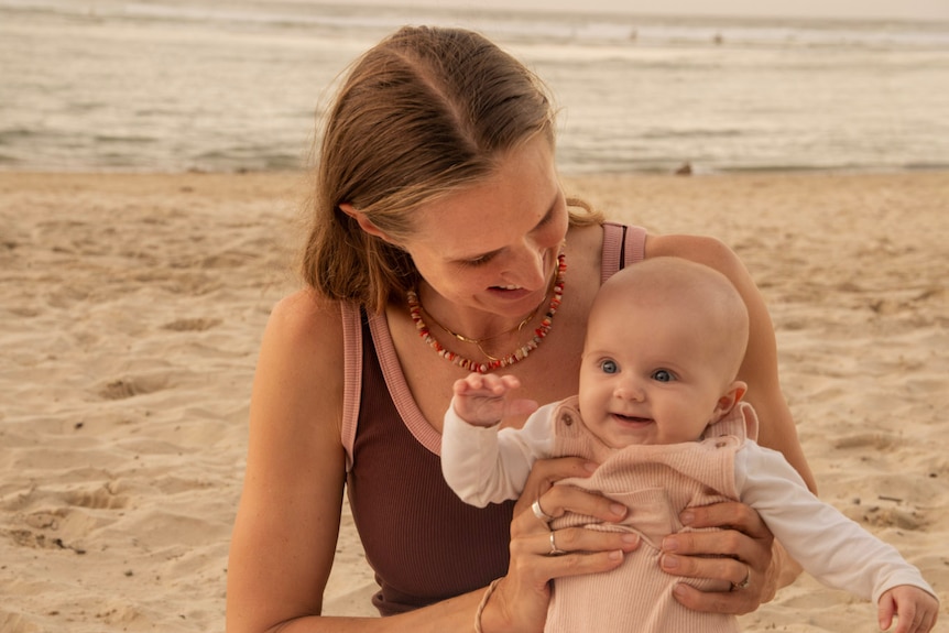 A woman looks at her baby and smiles while sitting on a beach.