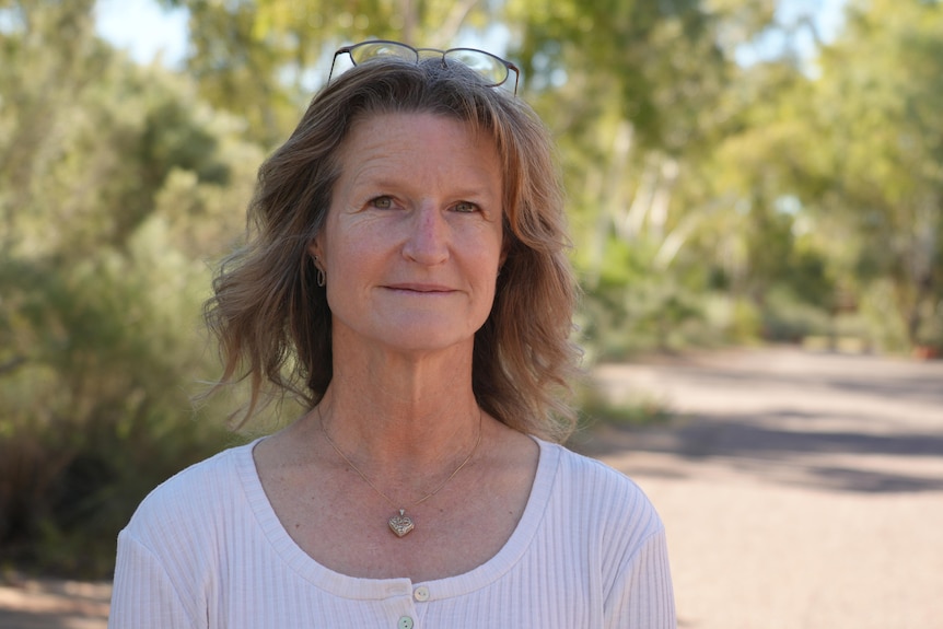 A woman wearing a white shirt and loveheart necklace smiles at the camera. There's trees in the background.