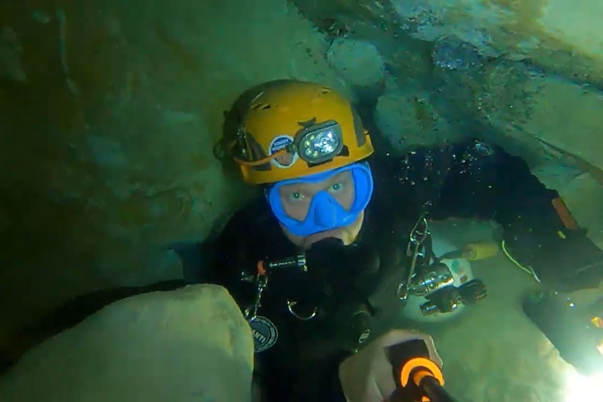 A cave diver wearing a yellow helmet, blue mask and black wetsuit squeezes between rocks.