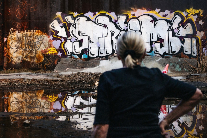 A woman stands with her arm on her hip, looking at huge storage tanks covered with graffiti.