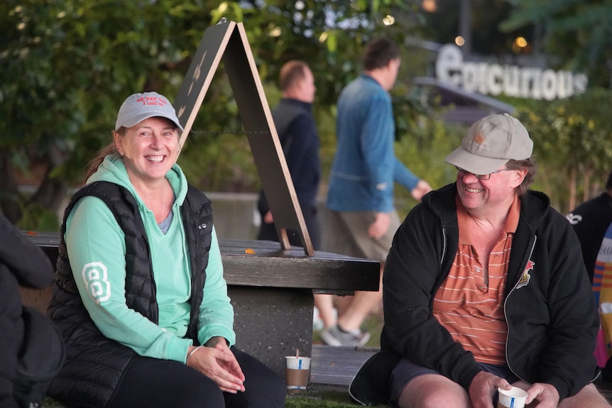 A woman and a man sit on a bench at a cafe laughing with two people walking in the background.