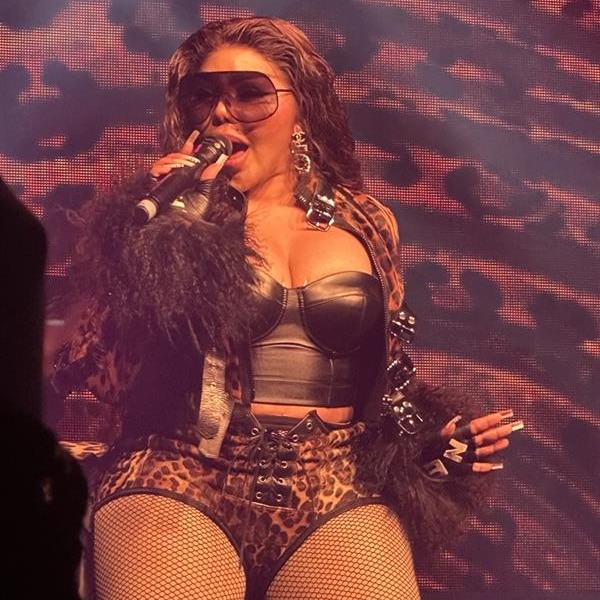 Lil' Kim holding mic in hand rapping on stage, wearing sunglasses and a leather bodysuit.