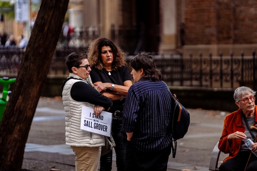 Three women huddle together, arms crossed. One holds an I stand with Sall Grover sign.