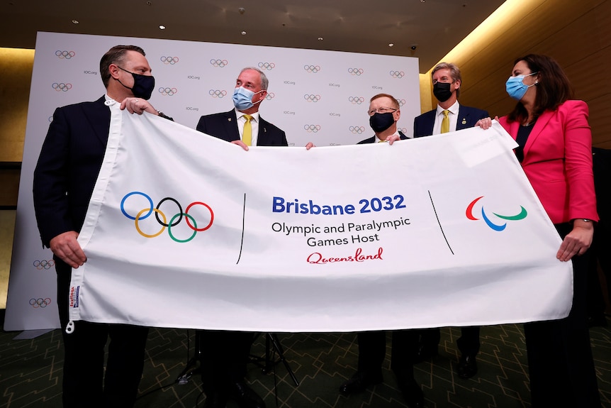 Four people, three men and one woman, holdingup a flag for Brisbane 2032 with the Olympics and Paralympics logo.