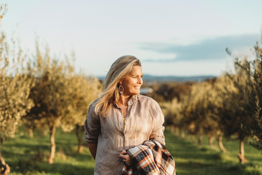 A blonde woman standing in a orchard in warm sunlight, smiling to her left.