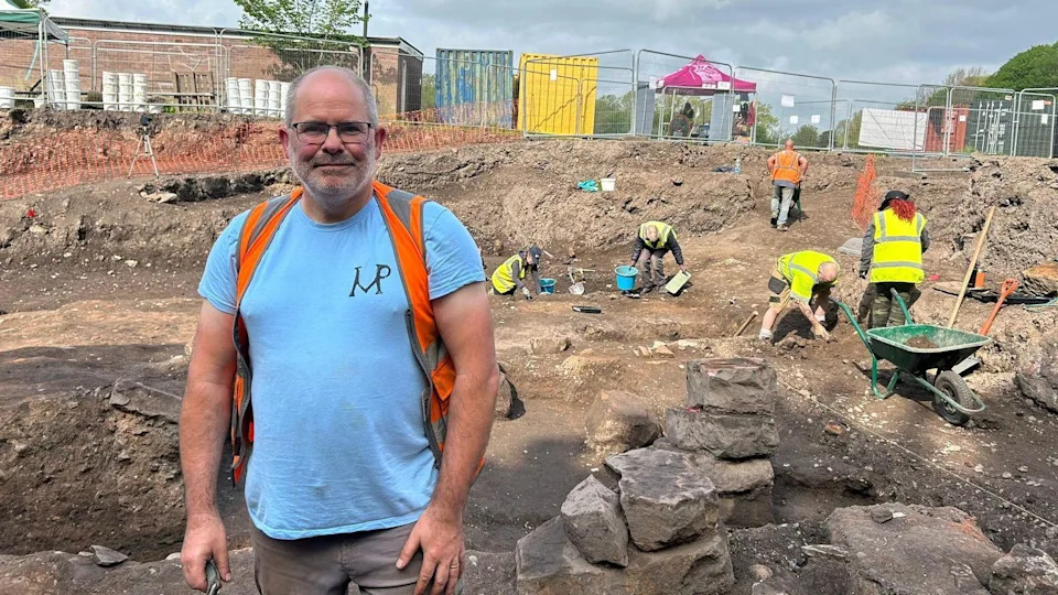 Frank Giecco has short grey hair at the sides. He is wearing glasses, a blue t-shirt, orange/grey rucksack, beige trousers. He is stood on the dig site with soil and rocks. There are volunteers with PPE on with tools and a green wheelbarrow. There is a temporary metal fence in the background a small brick building and large storage shipping containers.