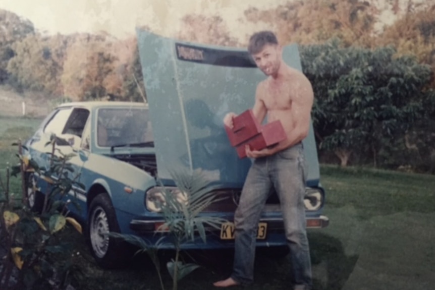 A young man in jeans and no shirt stands near a car with its bonnet raised.