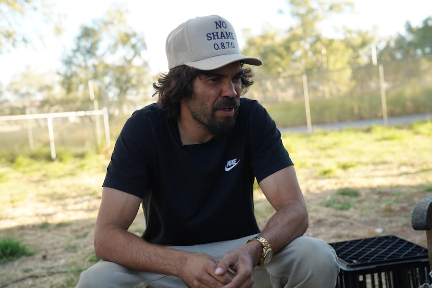 A young man in a baseball cap sits outside on a chair.