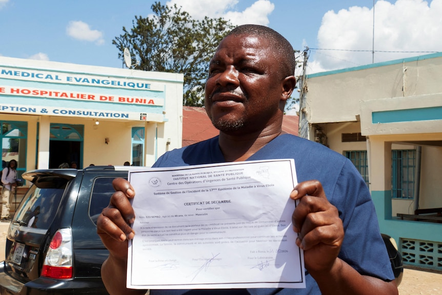 A man standing outside a medical clinic holding a certificate stating he has recovered.
