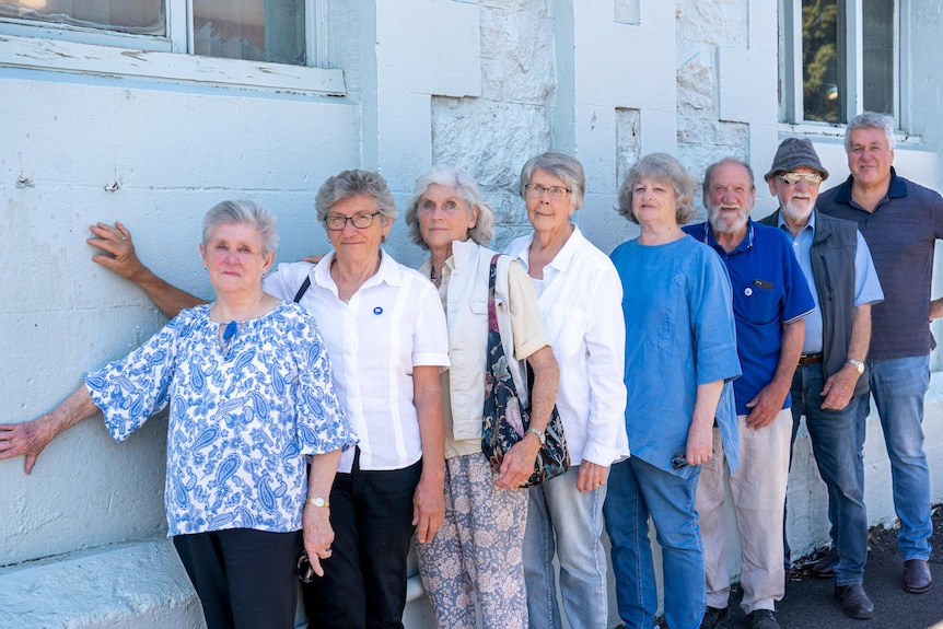 A group of people standing in a line in front of a pale blue wall, with some resting their hands on the wall.