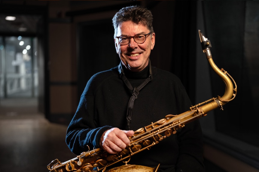 Man in a suit jacket and pants wears glasses, smiling, while holding a saxophone
