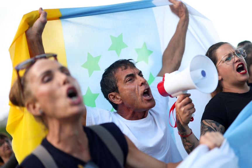 Three people stand, one with a megaphone, the other holding a flag over his head while protesting in Spain. 