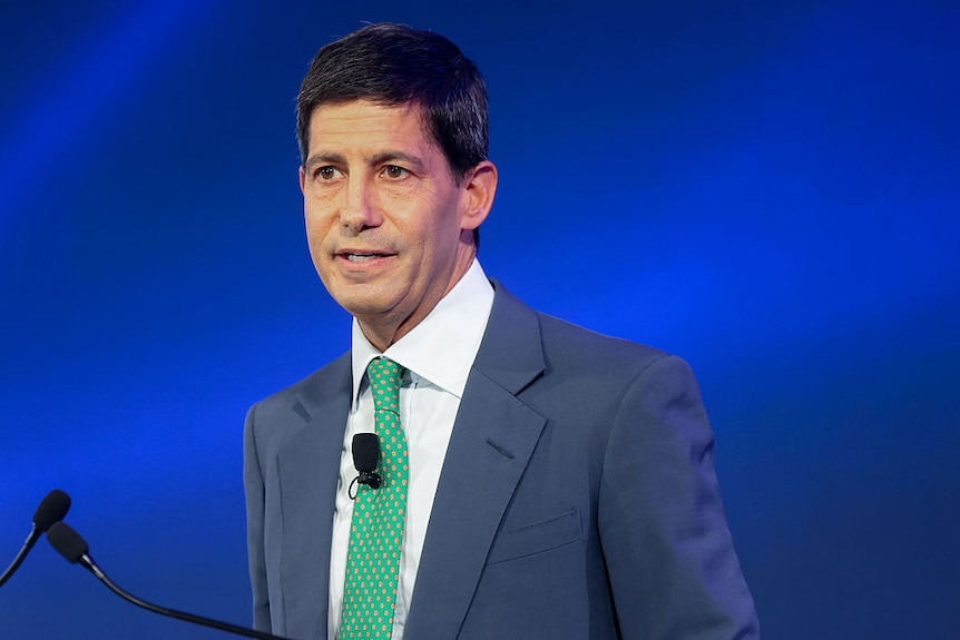 A photo of a man in his 40s with short black hair, a grey suit and brown eyes. He's wearing a green tie.