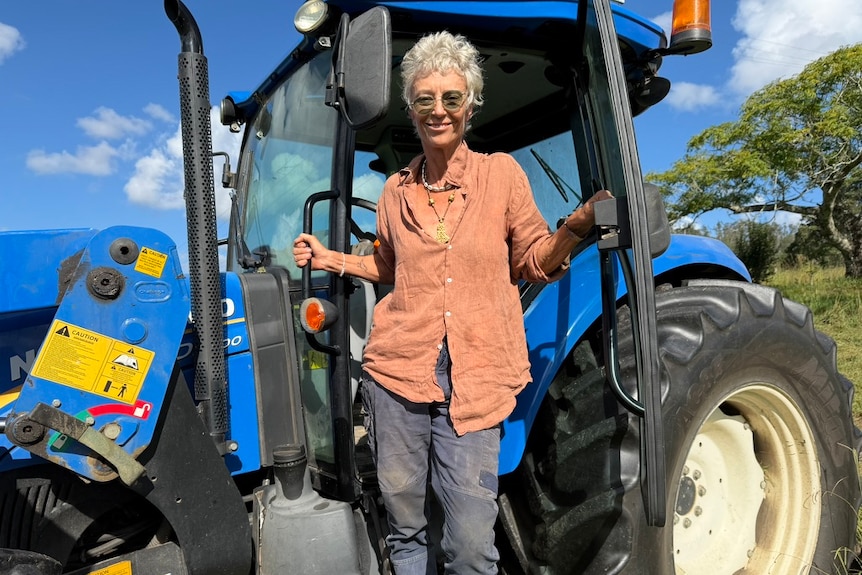 Woman with short grey hair wears glasses, long pants, buttoned blouse holding to the handles, standing on side of blue tractor