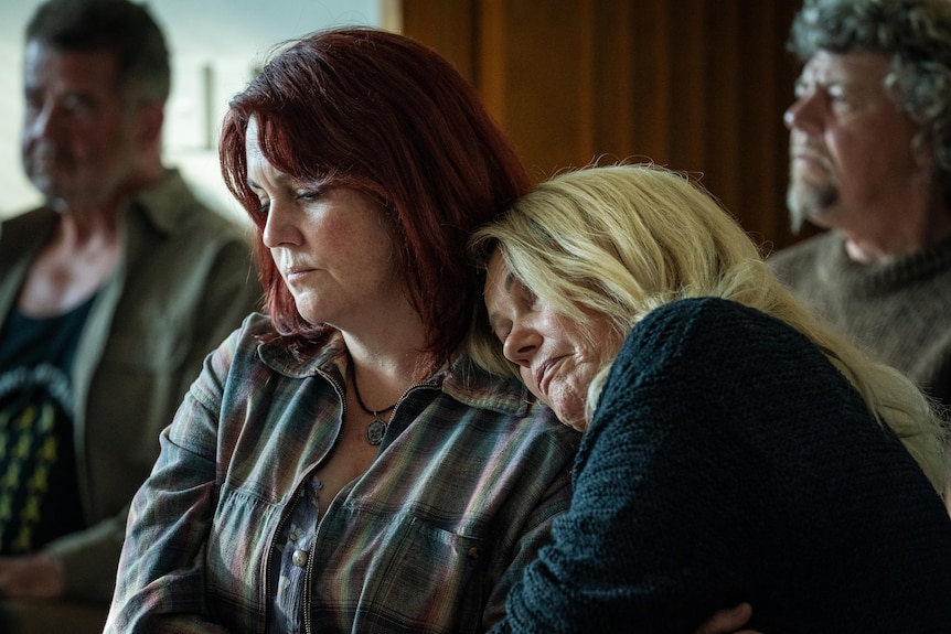 A woman with blonde hair rests her head on the shoulder of a woman with red hair.