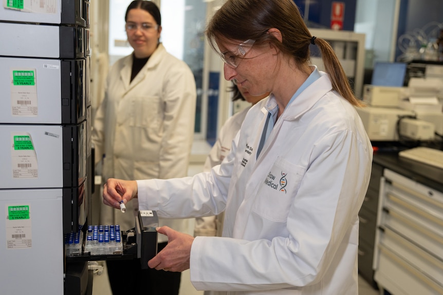 A woman wearing a lab coat looks at a small clear tube in a laboratory.