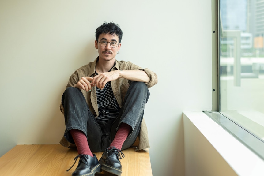A person with short hair, glasses and earrings, wearing a shirt and black jeans, sitting on a desk beside a window.