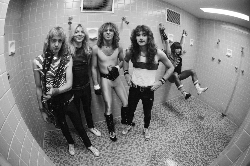 Iron Maiden pose with instruments in a tiled backstage shower room, wearing stage outfits, black and white photo