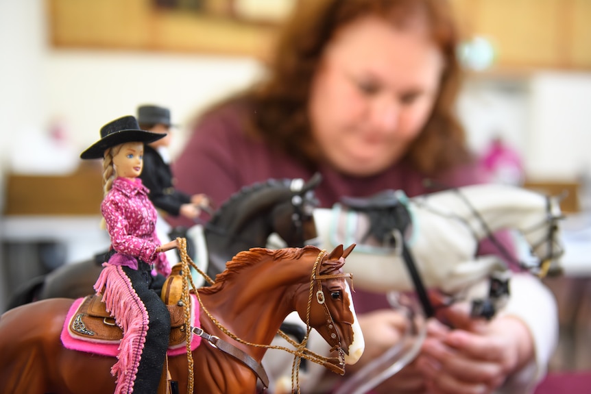 Brunette woman adding tack to model horses, barbie on brown horse salient