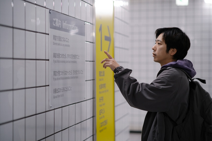 A still from a film showing a man pointing at a sign on a train station hallway wall.