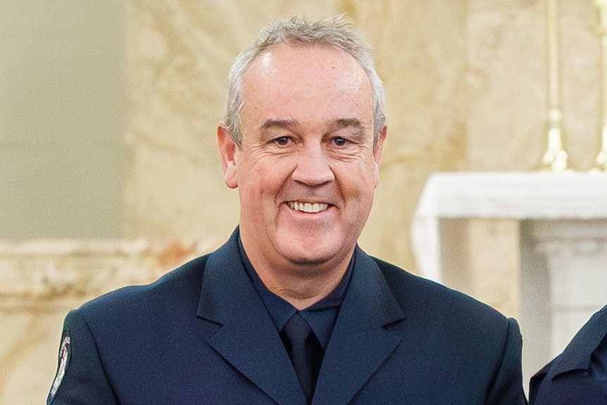 A man with grey hair standing, smiling while wearing a Victoria Police blazer and medals on his chest.