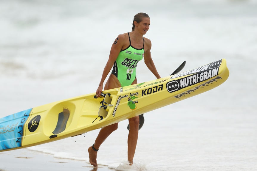 A woman in bright green swimmers walks along the beach holding a surf ski and paddle.