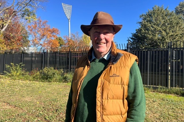 A man wearing an akubra stands outside the fence around an oval with tilted floodlights.