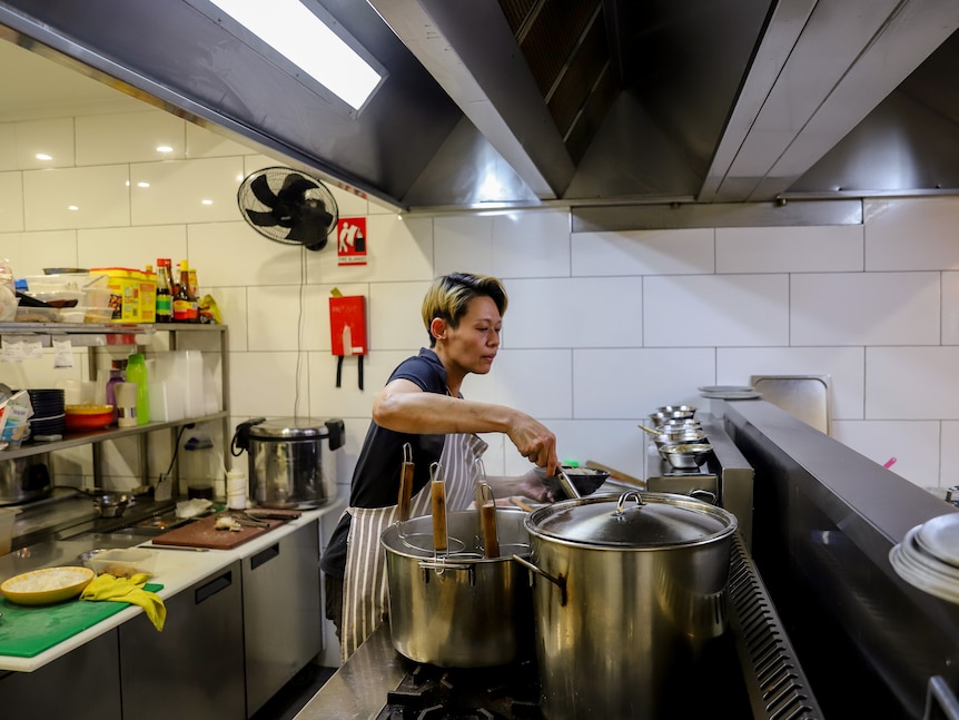 A woman with short cropped hair wearing an apron stands in front of stainless steel pots in a kitchen, she is stirring a spoon i