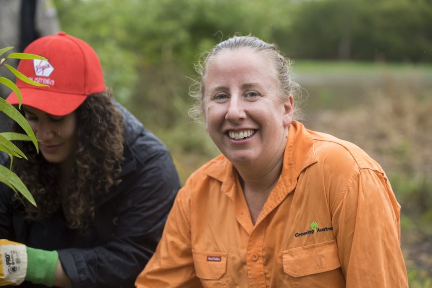 Lynise smiles at the camera, wears orange shirt with a logo, a woman in red cap, long hair looking at a plant beside her.