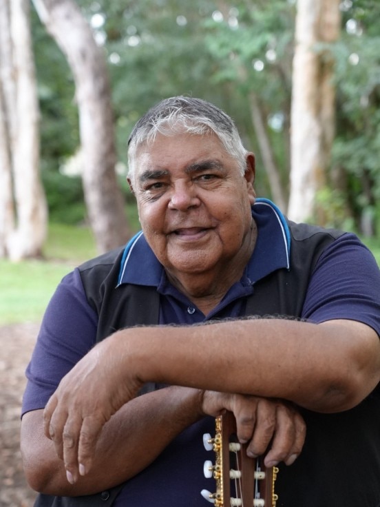 Uncle Roger Knox sits in the bush and rests his arm on his guitar.