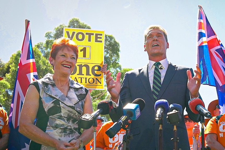 Cory Bernardi speaking at a podium with Pauline Hanson by his side and two Australian flags being waved in the background.