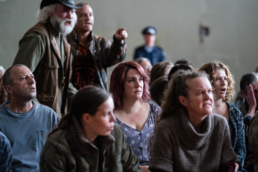 A group of angry and shocked people, with one standing and pointing, and a police officer in the background.