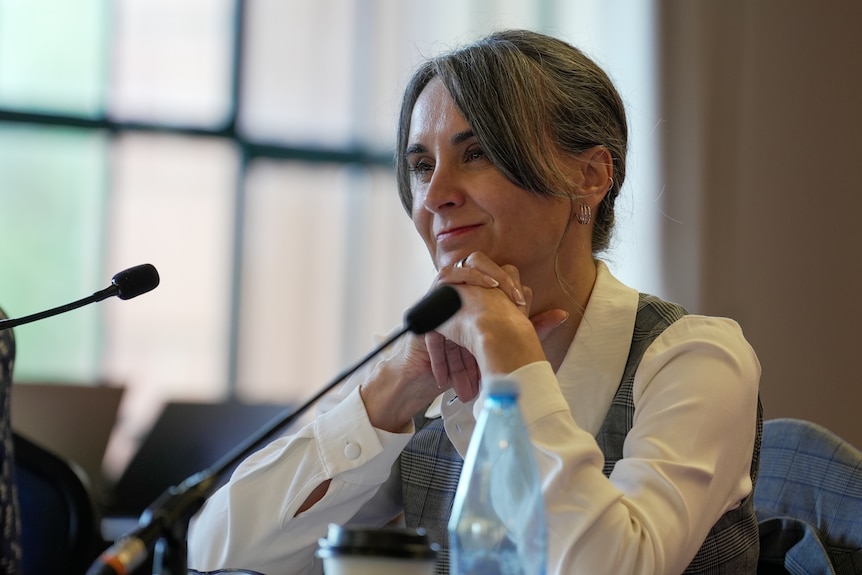 Woman in a white shirt and grey vest listening at a public hearing.