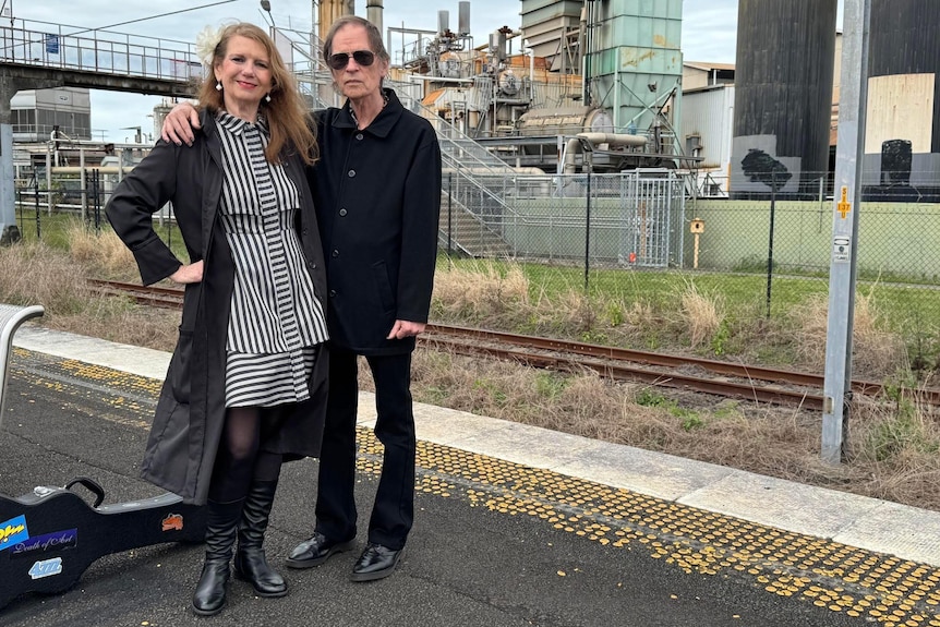 Mick Medew is dressed in black with sunbglasses on, with his arm around Ursula. They are at a train station.